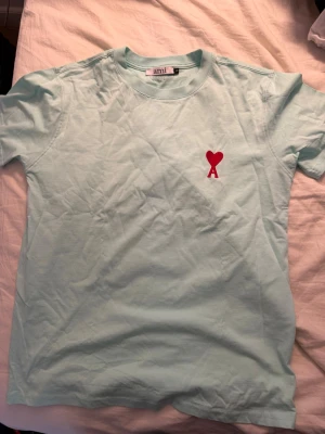 Ljusblå Ami Paris t-shirt med hjärtdetalj - Snygg ljusblå t-shirt från Ami Paris med klassisk passform och korta ärmar. På bröstet finns en röd broderad detalj med hjärta och bokstaven A. Tillverkad i mjuk bomull som känns skön mot huden. Perfekt för en clean och stilren look. Inte @kta men väldigt snygg ändå! 