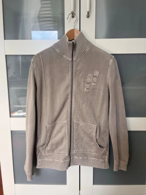 Beige ziptröja från Esprit - Snygg beige ziptröja från Esprit med hög krage och ribbade muddar. Tröjan har två stora fickor framtill och ett broderat mönster på bröstet. Tillverkad i mjuk bomullsblandning, perfekt för lager på lager och chill vardagsstil.