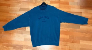 Blå stickad tröja från Nautica - Snygg blå stickad tröja från Nautica med rund halsringning och broderad text 'NAUTICA EST. 1983' på bröstet. Tröjan har långa ärmar och ribbade muddar vid ärmslut och nederkant. Perfekt för en avslappnad och stilren look.