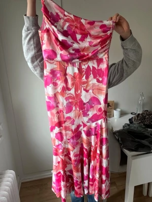 Blommig långklänning i rosa och rött - Säljer en långklänning med blommigt mönster i rosa, rött och vitt. Klänningen har en mjuk, böljande form och är ärmlös med en lätt draperad halsringning. Perfekt för sommarens alla tillfällen!