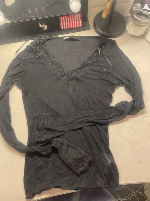 Svart långärmad topp med spets - Svart långärmad topp från Bik Bok med djup v-ringning dekorerad med spetsdetaljer. Tunn och mjuk i materialet, perfekt att styla med jeans eller kjol. Enkel och stilren design med en feminin touch tack vare spetsen vid halsen.
