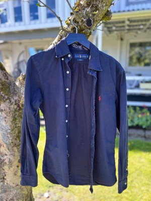 Mörkblå skjorta från Ralph Lauren - Snygg mörkblå skjorta från Ralph Lauren med klassisk krage och knappar framtill. Skjortan har lång ärm och den ikoniska röda loggan broderad på bröstet. Perfekt för en stilren look och passar till många olika outfits.