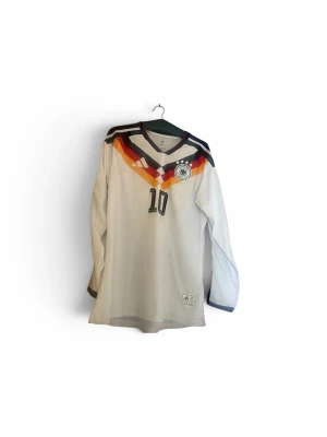Tyskland Musiala #10 Matchtröja – Långärmad 🇩🇪 - Säljer en riktigt clean Tyskland-tröja med Musiala #10 på ryggen. Långärmad modell med klassisk vit design och de ikoniska färgerna över bröstet. Perfekt för fotbollsfans, samlare eller till vardags.  * Spelare: Musiala * Nummer: 10 * Landslag: Tyskland * Märke: Adidas * Storlek: L * Skick: Väldigt fint skick * Långärmad modell * Climacool-material  Snygg detalj med patch på ärmen och autentisk look.  📩 Hör av dig vid intresse eller frågor!