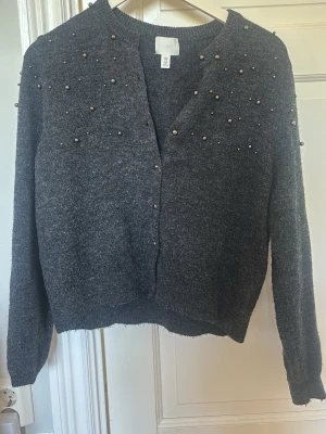 Mörkgrå kofta med pärlor från H&M - Passar även Xs och S. Mysig mörkgrå kofta från H&M med dekorativa pärlor på axlar och bröst. Koftan har lång ärm, rund halsringning och är stickad i ett mjukt material. Perfekt för lager-på-lager och en trendig look. 