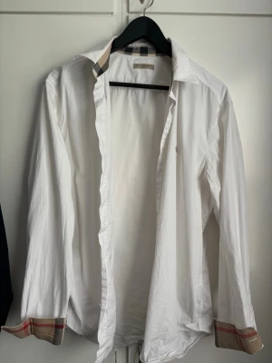  skjorta från Burberry Brit - Vit skjorta från Burberry storlek L med klassiska rutiga detaljer på krage och ärmslut. Skjortan har lång ärm och knappar framtill. Perfekt för en stilren och tidlös look.