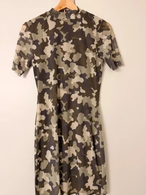 Kort camoklänning med kort ärm - Cool kort klänning med camouflage-mönster i gröna och beige toner. Klänningen har korta ärmar och en rund halsringning. Perfekt för dig som vill sticka ut med en edgy stil. Materialet känns mjukt och stretchigt, troligen polyesterblandning.