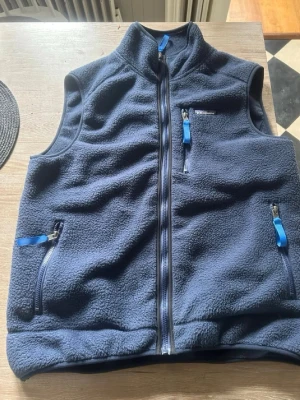 Mörkblå fleeceväst från Patagonia - Mysig mörkblå fleeceväst från Patagonia med hög krage och tre fickor med dragkedja. Västen har blå detaljer på dragkedjorna och är perfekt för lager-på-lager. Klassisk och enkel design som funkar till det mesta.