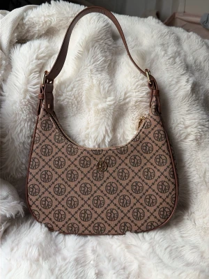 Axelremsväska - Snygg beige handväska med brunt monogramtryck över hela väskan och guldfärgad logga framtill. Väskan har en böjd form, justerbart axelband i brunt konstläder och dragkedja. Köpt för 200kr och aldrig använd🫶