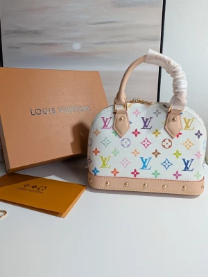 Iouis Vuitton BB Murakami edition 🪷 - En dröm i färg och form Denna ljuvliga handväska utstrålar en tidlös charm med sin mjuka, kupolformade silhuett som påminner om klassisk elegans. Den står stadigt på en vacker bas, redo att bära dina mest värdefulla ägodelar genom livets alla små och stora ögonblick.Väskan omfamnas av två handtag i naturligt, ljusbeige läder som med tiden kommer att bära spår av dina resor och äventyr.