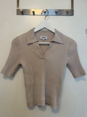 Beige ribbad topp från ONLY - Snygg beige ribbad topp från ONLY med v-ringning och krage. Toppen har korta ärmar och en figurnära passform. Inga fler bilder skickas då det ligger i flyttlåda.