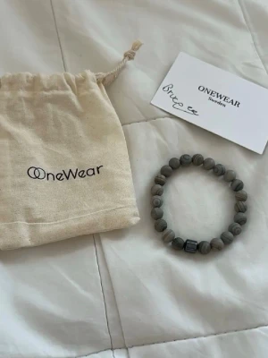 Grått pärlarmband från OneWear - Stilrent armband från OneWear med runda grå pärlor och en mörkare fyrkantig detalj. Armbandet har en elastisk tråd och levereras med en tygpåse med logga. Perfekt accessoar för att lyfta din outfit.