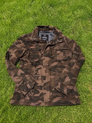 Camo vindjacka från Superdry - Snygg vindjacka från Superdry i klassiskt militärinspirerat camouflagemönster. Jackan har fyra fickor framtill, hög krage och knappar vid ärmsluten. Perfekt för dig som gillar streetstyle och vill sticka ut med en cool look.