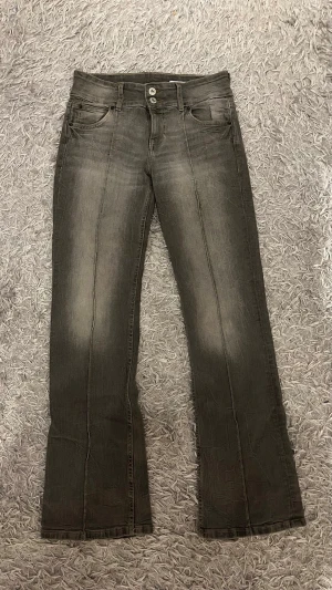 Grå bootcut jeans med slitningar - Snygga grå bootcut jeans low waist. Jeansen har dubbla knappar i midjan och subtila ljusare partier framtill och baktill för en cool vintagekänsla. Perfekta för dig som gillar en avslappnad men trendig stil.