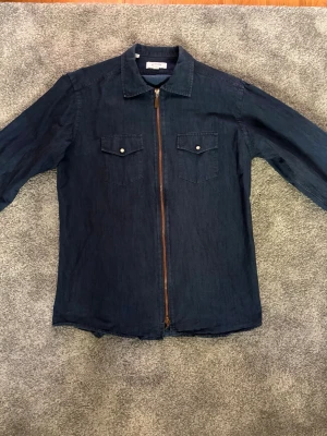 Mörkblå overshirt från Eton - Snygg mörkblå overshirt från Eton i denimliknande material. Modellen har dragkedja framtill, två bröstfickor med lock och knapp samt klassisk krage. Perfekt lager-på-lager-plagg med lång ärm och stilren look. Perfekt inför sommaren. Den är i bra skick. Hör av er vid minsta fundering!