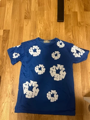 Denim tears t shirt blue  - This blue denim tears short-sleeved t-shirt features a distinctive white abstract circular pattern, composed of interlocking figures, distributed across the front, back, and sleeves. The fabric has a smooth texture, suggesting a comfortable and versatile wear for casual or athletic activities.  . #bluetshirt #abstractprint #graphictee #casualwear #athleticwear #secondhandfashion #unisexfashion #circularpattern #whiteprint #shortleeve