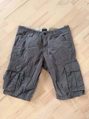 Grå cargoshorts med fickor - Säljer ett par grå cargoshorts med flera stora fickor på sidorna och baktill. Shortsen har normal passform, knapp och dragkedja framtill samt bälteshällor. Perfekta för en avslappnad stil under sommaren.