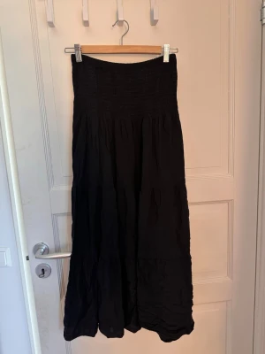Svart långkjol med smock i midjan - Säljer en svart långkjol med smock i midjan och volangdetaljer. Kjolen har en luftig och flowy passform, perfekt för dig som gillar en avslappnad men ändå stilren look. Materialet känns mjukt och bekvämt mot huden.