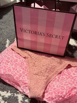 Rosa spetsstring från Victoria's Secret - Supersnygga rosa stringtrosor från Victoria's Secret i mjuk spets. Trosorna har en feminin design med blommigt spetsmönster och låg midja. Perfekta för dig som gillar bekväma och stilrena underkläder med en touch av lyx.