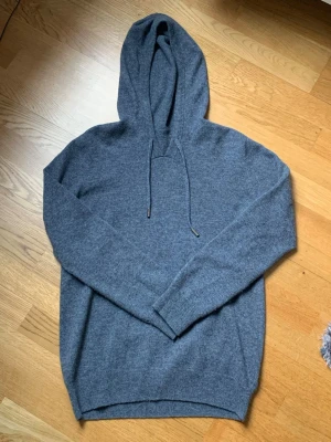 Mörkgrå merinoull hoddie - En stilren mörkgrå hoodie med huva. Tillverkad i Kashmir och merinoull som är ett mjukt och skönt tyg, perfekt för en avslappnad look. Långa ärmar och ribbade muddar ger en snygg passform. Passar perfekt till jeans eller joggers. Måste tyvärr sälja denna nu pga att den blivit för liten. 