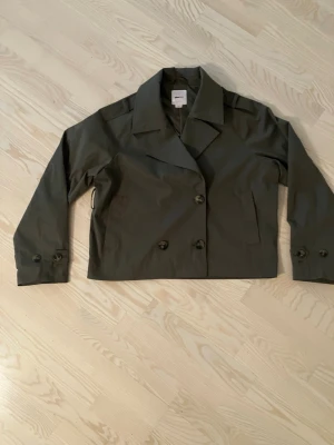 Olivgrön kort rock med bred krage från Gina Tricot - Stilren olivgrön rock med bred krage och dubbelknäppning framtill. Modellen är kortare och har knappar vid ärmsluten för extra detalj. Perfekt för dig som gillar minimalistisk och modern stil.