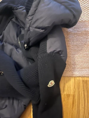 Momcler tricot Cardigan - Snygg svart dunjacka från Moncler med stickade partier och klassisk logga på ärmen. Jackan har dragkedja framtill och en mysig huva. Perfekt för kalla dagar och ger en stilren look.