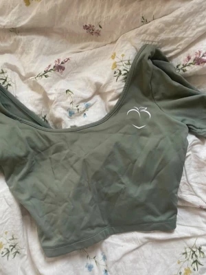 Grön croppad topp från Peach - Snygg ljusgrön croppad topp från Peach med omlottdetalj i ryggen och korta ärmar. Toppen har en stilren design med en liten vit logga framtill och är tillverkad i mjukt, stretchigt material. Fint skick! 