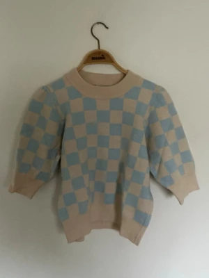 Stickad tröja med rutigt mönster - Snygg stickad tröja med ljusblått och beige rutmönster. Tröjan har puffiga trekvartsärmar, bred ribbad mudd vid hals, ärmslut och nederkant. Perfekt för dig som gillar en trendig och lekfull stil.
