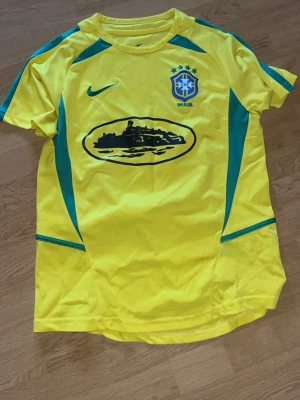 Corteiz brasilien tröja - Gul fotbollströja från Nike med Brasilien-emblem på bröstet och gröna detaljer längs sidorna. Tröjan har rund hals och korta ärmar, samt ett svart tryck på magen. Perfekt för fotbollsträning eller att bära som supporter.