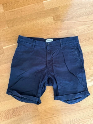 Mörkblå shorts från These Glory Days - Snygga mörkblå shorts från These Glory Days med klassisk passform och uppvikta benslut. Shortsen har bälteshällor, sidofickor och två bakfickor. Tillverkade i mjuk bomull som känns skön mot huden. Perfekta för varma dagar.