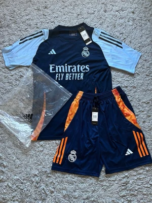 Real Madrid träningsset Adidas [FAST PRIS] - Snyggt Real Madrid träningsset från Adidas med tröja och shorts. Tröjan är mörkblå med ljusblå ärmar, vita loggor och orange detaljer. Shortsen är mörkblå med orange ränder och Real Madrid-emblem. Tillverkat i lätt och ventilerande material.