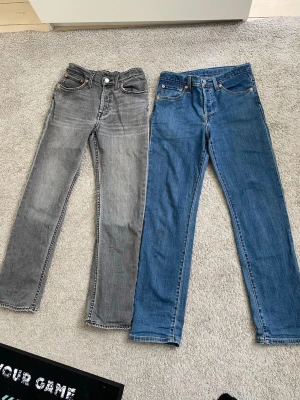 Två par Levis 501  jeans i olika färger - Säljer två par raka jeans, ett par i klassisk blå denim och ett par i tvättad grå. Båda har fem fickor, knappgylf och kontrastsömmar. Perfekta för en avslappnad och trendig look. Materialet är bomullsdenim. 