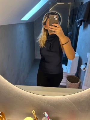 Marinblå  ärmlös halterneck topp - Snygg svart ärmlös halterneck topp med hög hals och slät yta. Toppen har en elegant och stilren design som passar perfekt till många olika outfits. Materialet ser ut att vara mjukt och har ett lätt glansigt utseende.