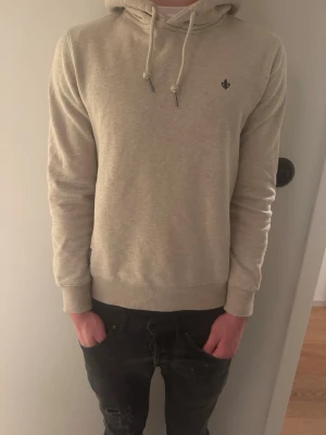 Beige hoodie från Morris Stockholm - Snygg beige hoodie från Morris Stockholm med broderad logga på bröstet. Klassisk modell med huva och dragsnören samt ribbade muddar vid ärmslut och nederkant. Perfekt för en avslappnad och stilren look.