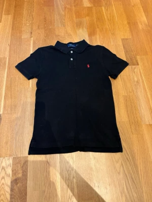 Svart polotopp från Polo Ralph Lauren - Klassisk svart polotopp från Polo Ralph Lauren med krage och knappar framtill. Ikonisk röd logga broderad på bröstet. Perfekt för en clean och stilren look. Gjord i mjuk bomull som känns skön mot huden.