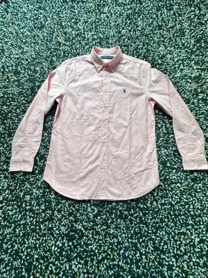 Ljusrosa skjorta från Ralph Lauren - Klassisk ljusrosa skjorta från Ralph Lauren med button-down krage och broderad logga på bröstet. Skjortan har långa ärmar, knappar framtill och är tillverkad i bomull. Perfekt för en stilren och avslappnad look. Helt ny utan prislapp! 