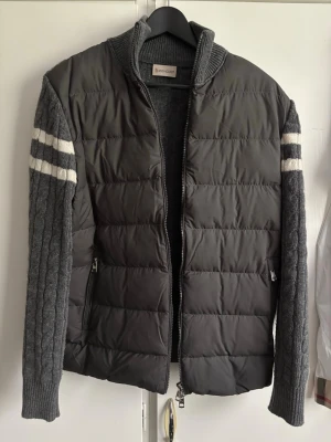  Moncler cardigan - Snygg grå cardiganjacka från Moncler storlek S-M med vadderad framsida och stickade ärmar samt rygg. Ärmarna har vita ränder och kabelstickat mönster. Jackan har hög krage, dragkedja och två fickor med dragkedja framtill.