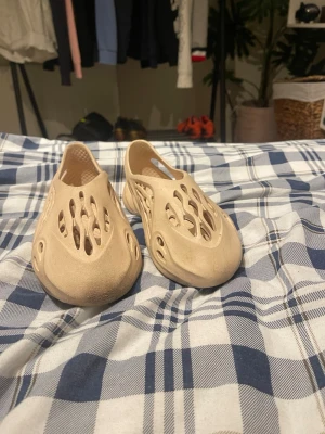 Beige Yeezy Foam Runner tofflor - Säljer ett par beige Yeezy Foam Runner tofflor med futuristisk design och många ventilationshål. Tillverkade i lätt och bekväm EVA-skum som formar sig efter foten. Perfekta för dig som gillar streetwear och vill ha något unikt på fötterna.