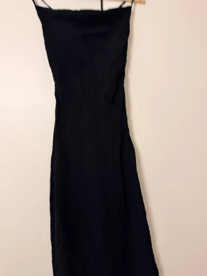 Svart långklänning från Gina Tricot - Stilren svart långklänning från Gina Tricot med smala axelband och öppen rygg med snörning. Klänningen har en elegant, figurnära passform och är tillverkad i ett lätt, något glansigt material. Perfekt för dig som gillar minimalistisk och trendig stil.