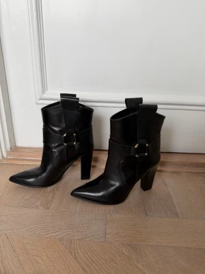 Svarta cowboy boots med klack - Snygga svarta cowboy boots i skinn med spetsig tå och bred klack. Bootsens design har dekorativa remmar med metallspännen och höga skaft med dragflikar. Inköpta på Marzio i Stockholm