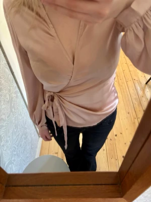Ljusrosa omlottblus med knytband - Snygg ljusrosa omlottblus i glansigt material med långa ärmar och knytband i midjan. Blusen har v-ringning och ballongärmar med resår vid ärmslut. Perfekt för en chic och trendig look.