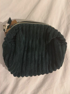 Mörkgrön necessär - Säljer en mörkgrön necessär med ribbad struktur och dragkedja i guld. Väskan är mjuk och gjord i frottéliknande material, perfekt för att förvara smink eller småprylar. Enkel och stilren design som sticker ut i väskan eller på hyllan.