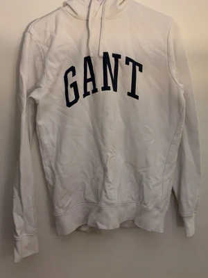 Vit hoodie från GANT med tryck - Vit hoodie från GANT med stort svart tryck på bröstet. Klassisk modell med huva och dragsnören, ribbade muddar och lång ärm. Perfekt för en avslappnad och cool look. Materialet är mjukt och bekvämt.