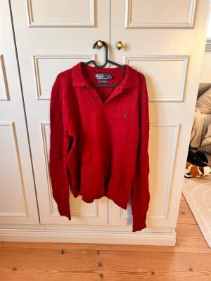 Röd tröja med krage från Polo Ralph Lauren - Röd långärmad tröja från Polo Ralph Lauren med klassisk krage och knappar vid halsen. Tröjan har det ikoniska blå broderade logotypen på bröstet och är tillverkad i ett mjukt stickat ull material. Perfekt för dig som gillar stilren och tidlös design.     Den här tröjan kan diskuteras pris mellan 700kr uppåt för att den är 100% ull