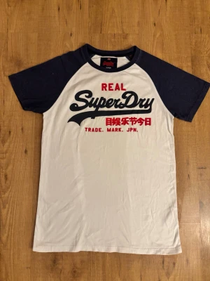Superdry vit och marinblå t-shirt - Snygg t-shirt från Superdry med vit framsida och marinblå ärmar. Stor logga och japansk text på bröstet i svart och rött. Klassisk rund halsringning och kontrastfärgade raglanärmar. Perfekt för en avslappnad och trendig stil.
