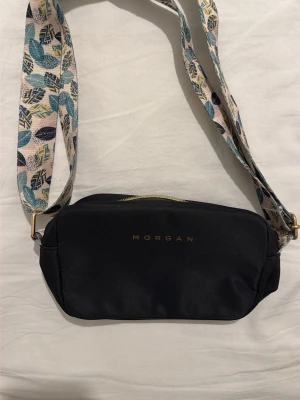 Svart axelväska från Morgan - Snygg svart axelväska från Morgan med guldfärgad dragkedja och ett brett, mönstrat axelband i vitt, blått och gult. Väskan har en enkel, stilren design med diskret logga framtill och är perfekt för att bära det viktigaste. Materialet känns tåligt och bandet ger en cool touch.