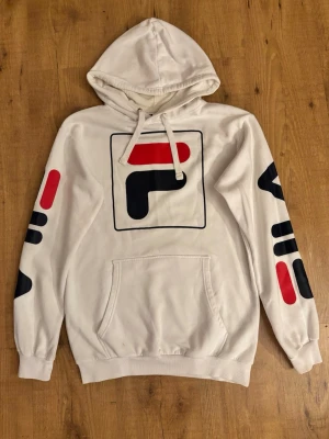 Vit hoodie från FILA med tryck - Snygg vit hoodie från FILA med stor logga på bröstet och tryck i svart och rött på båda ärmarna. Klassisk känguruficka framtill och spännbar huva. Perfekt för en sportig och avslappnad stil.