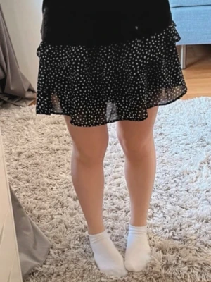 Svart kortkjol med vita prickar - Söt blå kortkjol med vita prickar och volangkant. Kjolen har en luftig och lätt känsla, perfekt för dig som gillar en lekfull och trendig stil. Passar till både sneakers och boots för en cool vardagslook.