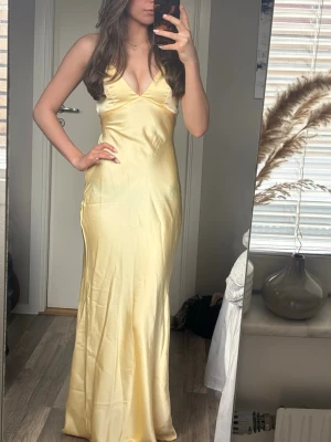 Ljusgul långklänning med v-ringning - Elegant ljusgul långklänning med v-ringad hals och ärmlös design. Klänningen har en slank passform och är tillverkad i ett glansigt material som ger en lyxig känsla. Perfekt för dig som vill sticka ut med en stilren och modern look. Med öppen rygg med knytning