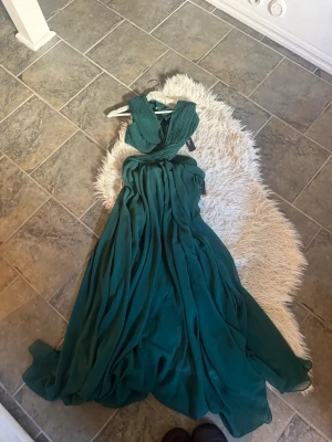 Grön långklänning med öppen rygg - Säljer en elegant grön långklänning med plisserad överdel och öppen rygg. Klänningen har ett flytande, svepande tyg och breda axelband. Perfekt för bal eller festliga tillfällen. Dragkedja i ryggen för enkel påklädning.