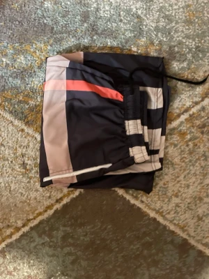 Randiga badbyxor med dragsko - Säljer ett par snygga badbyxor med breda svarta, beige och röda ränder. De har elastisk midja med svart dragsko och sidofickor. Materialet är snabbtorkande polyester, perfekt för stranden eller poolen.